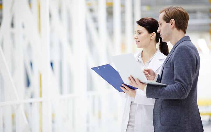 A man and a woman in a manufacturing plant. The man is holding a laptop and the woman is holding a clipboard.