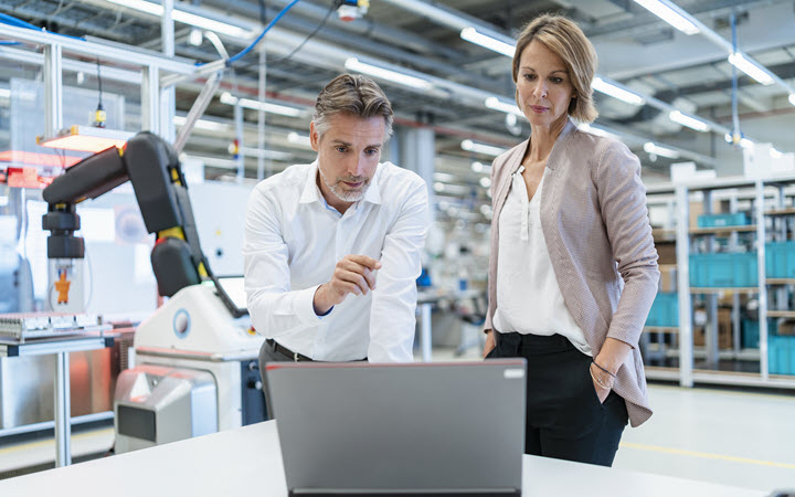 A man and a woman in a manufacturing plant working on a laptop screen. The man is pointing at the screen and the woman is looking at the screen.