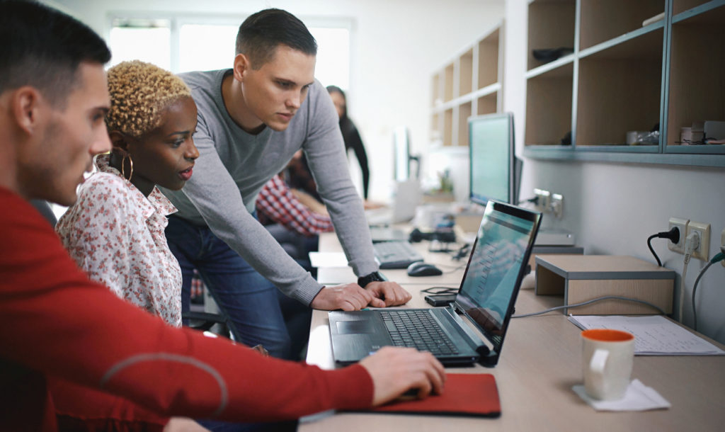 Application developers at work. Closeup of group of application developers discussing about certain code for an application. There's black woman two caucasian men gathered around a desktop computer. They are late 20's, mixed race, dressed smart casually. Blurry people in background, also released.