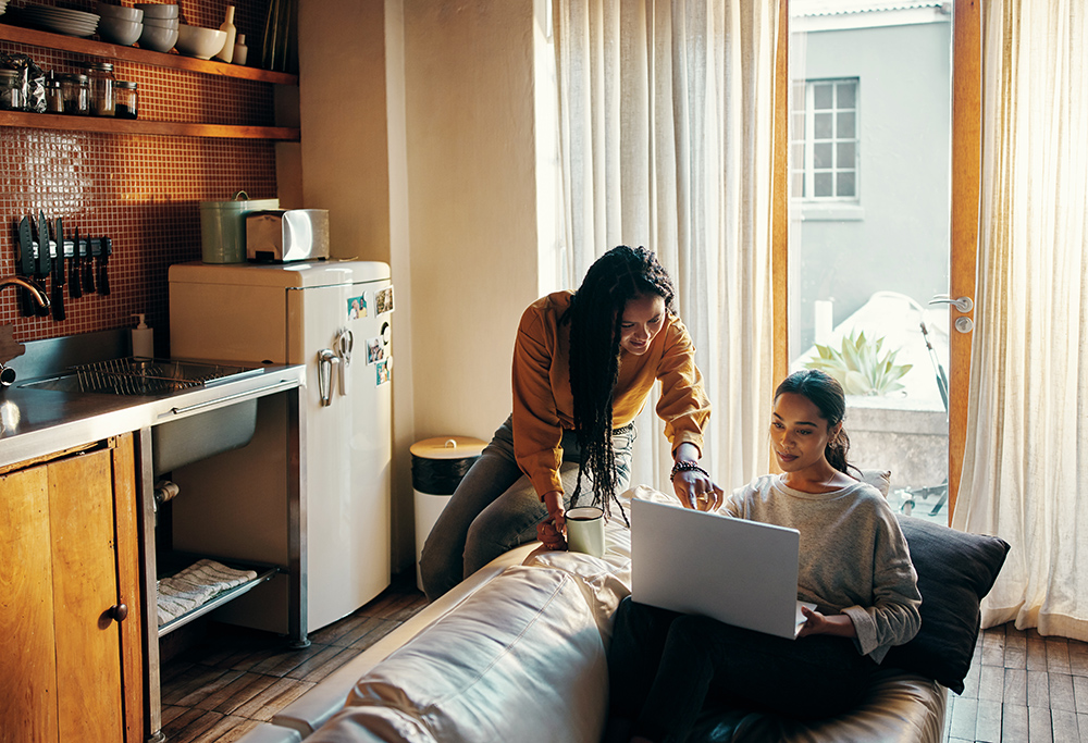 Shot of two attractive young women drinking coffee and using a laptop while relaxing together at home over the weekend