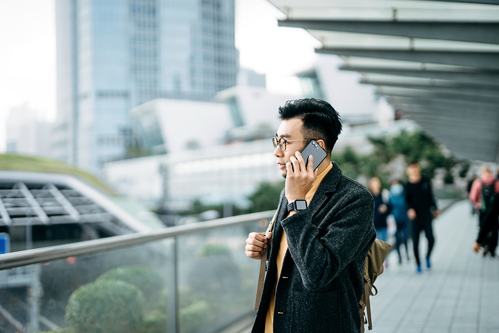 Portrait of young urban Asian businessman talking on smartphone and is carrying backpack on one shoulder while commuting in the city