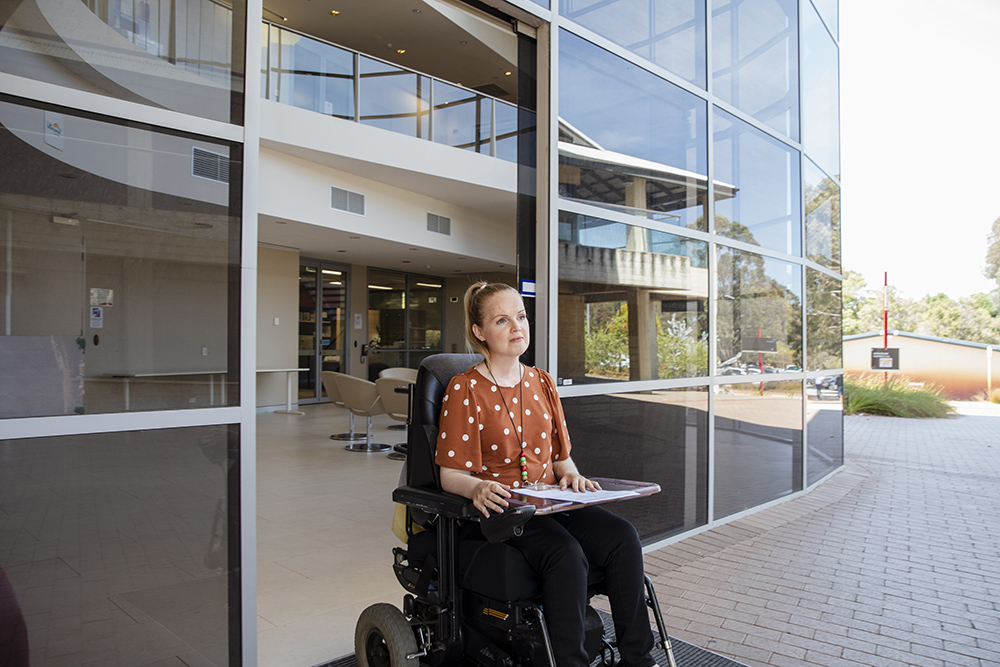 A front view of a woman leaving her office in her electric wheelchair take some paperwork to a colleague.