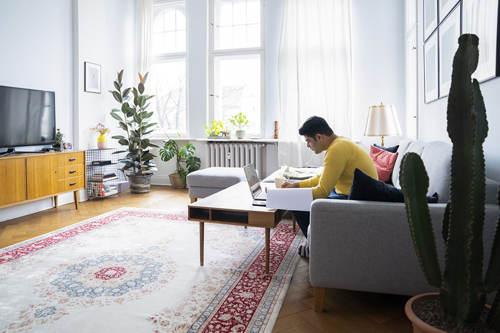 Side view of man using laptop at coffee table while sitting on sofa. Full length of male is financial planning at home. He is in living room.