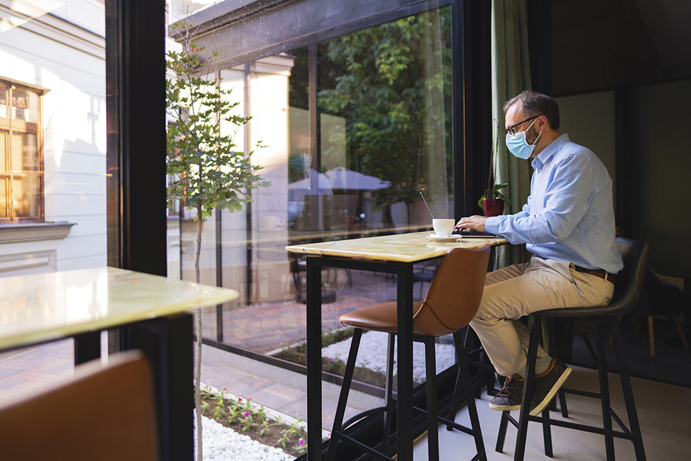 Mature Caucasian businessman with a protective face mask sitting at the coffee place and working on his laptop.