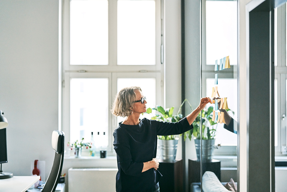 Businesswoman looking at adhesive notes stuck on glass wall. Mature female business professional is planning in office. She is in smart casuals.