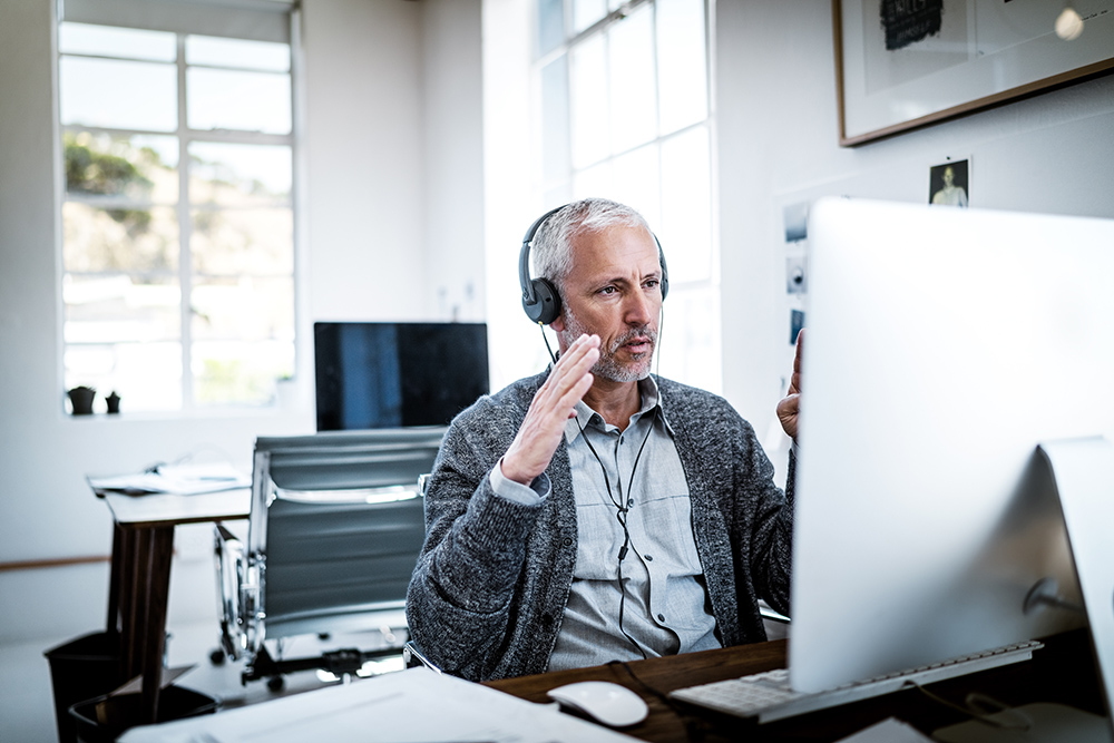 Confident businessman video conferencing on computer. Male professional is using headphones while sitting at desk. He is in brightly lit office.