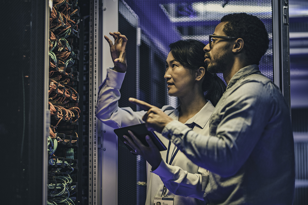 Side view of a female Asian IT engineer explaining network connections in a server to a novice African-American technician.