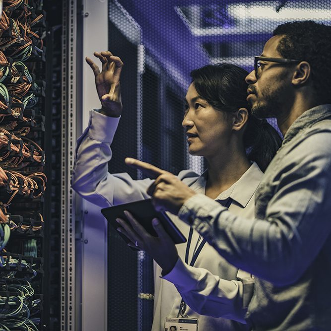 Side view of a female Asian IT engineer explaining network connections in a server to a novice African-American technician.